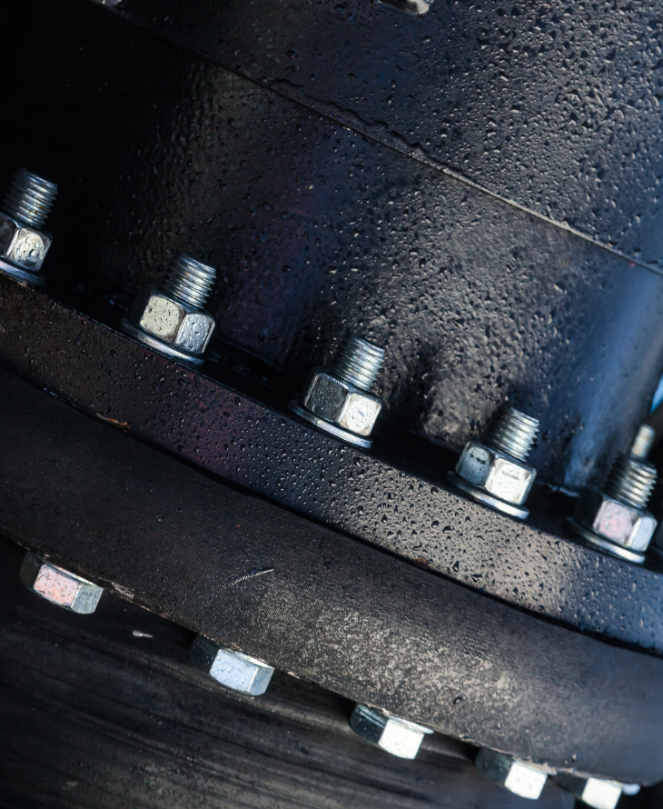 Close-up of a black metal flange secured with silver bolts and nuts, covered in water droplets.