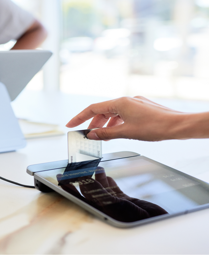  A hand inserts a credit card into a card reader on a counter with a blurred person and tablet in the background.