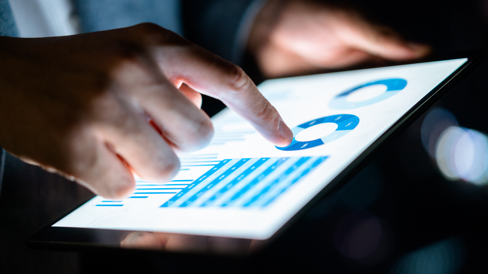 Close-up of hands tapping a tablet screen displaying blue pie charts and bar graphs.