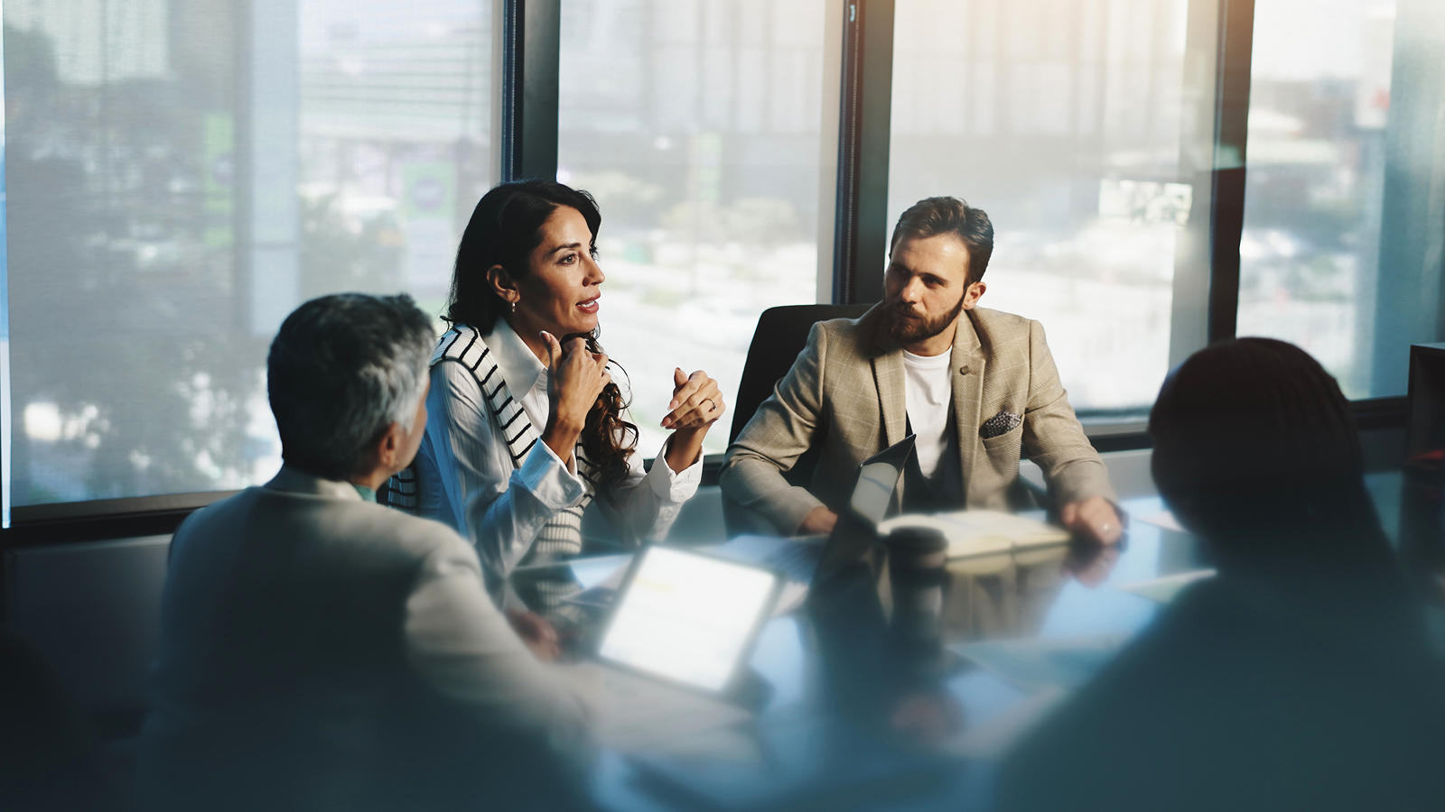 Small business meeting around a conference table with laptops and documents, city view through large windows.