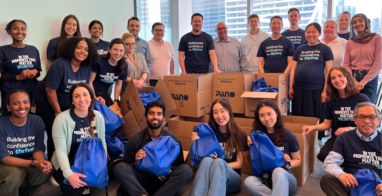 A group of volunteers stands in an office space, surrounded by boxes and bags, all wearing matching blue shirts that say “Building the confidence to thrive”, working on community service projects.