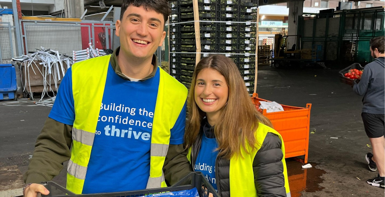 Two individuals with bright yellow vests are shown holding a tray of vegetables in a food distribution center, exemplifying volunteer work supporting food security.
