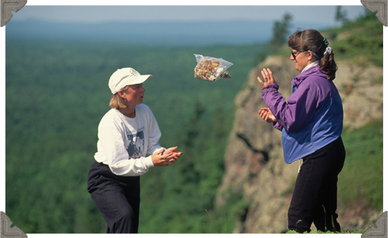 a picture of two woman throwing and catching a bag on a hill