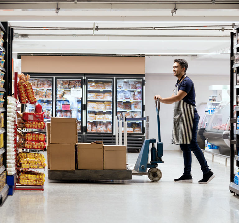 A supermarket worker uses a hand truck to transport boxes down an aisle, flanked by shelves and refrigerated display cases.