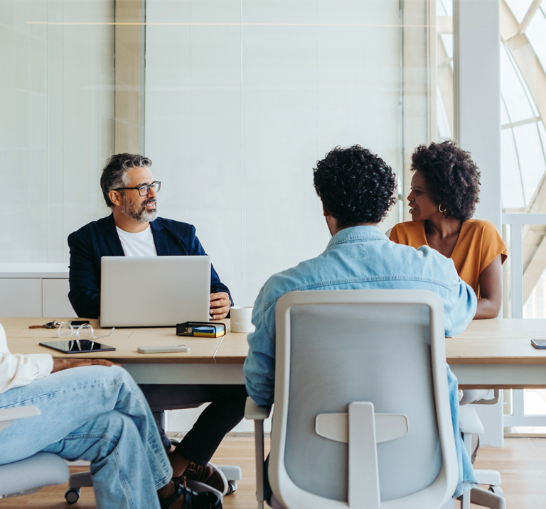 A diverse group sits around a modern conference table, engaged in discussion, with a laptop and documents displayed.