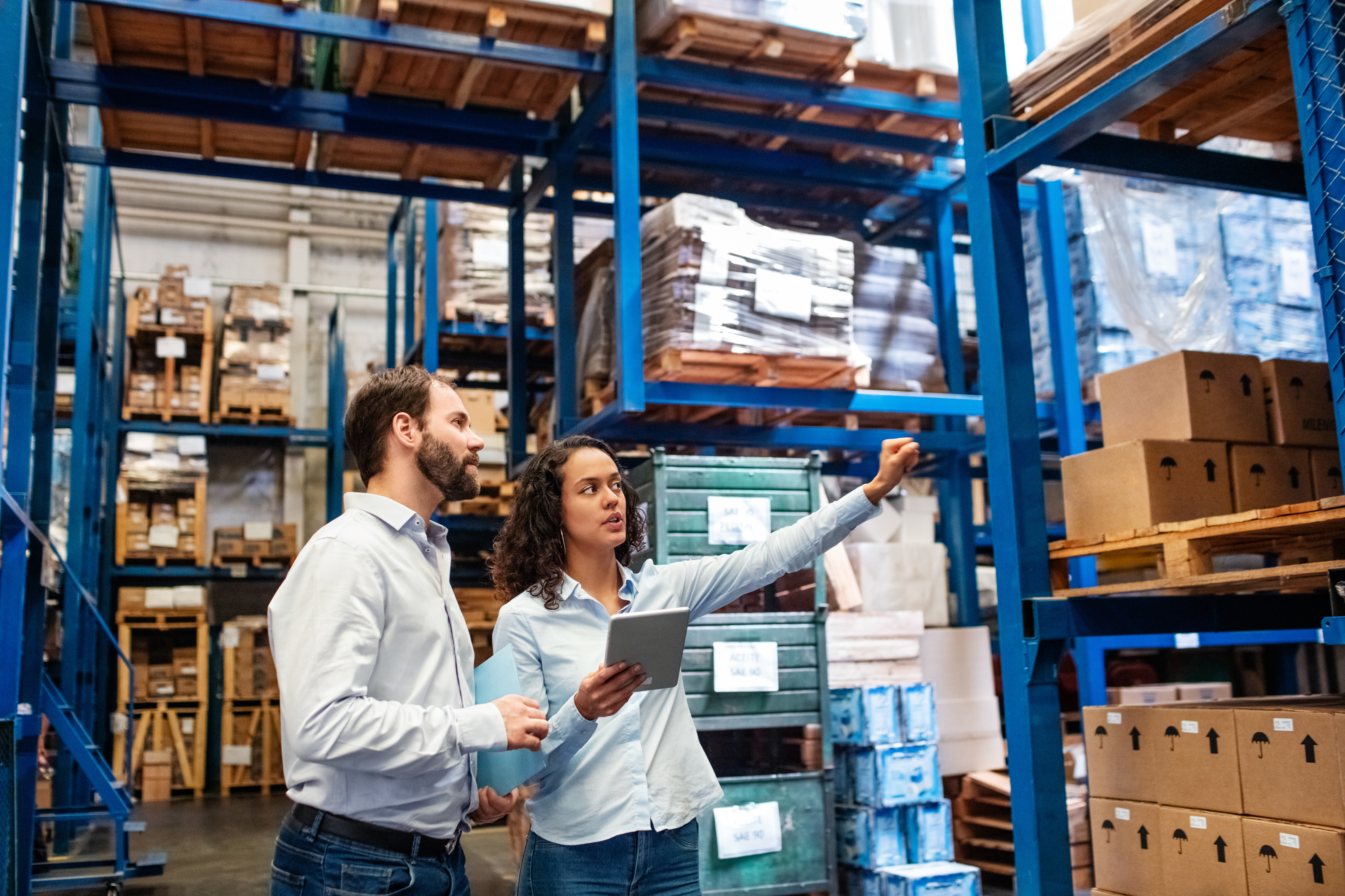 Two people in business attire stand in a warehouse; one holds a tablet while the other points toward shelves stacked with boxes and pallets.