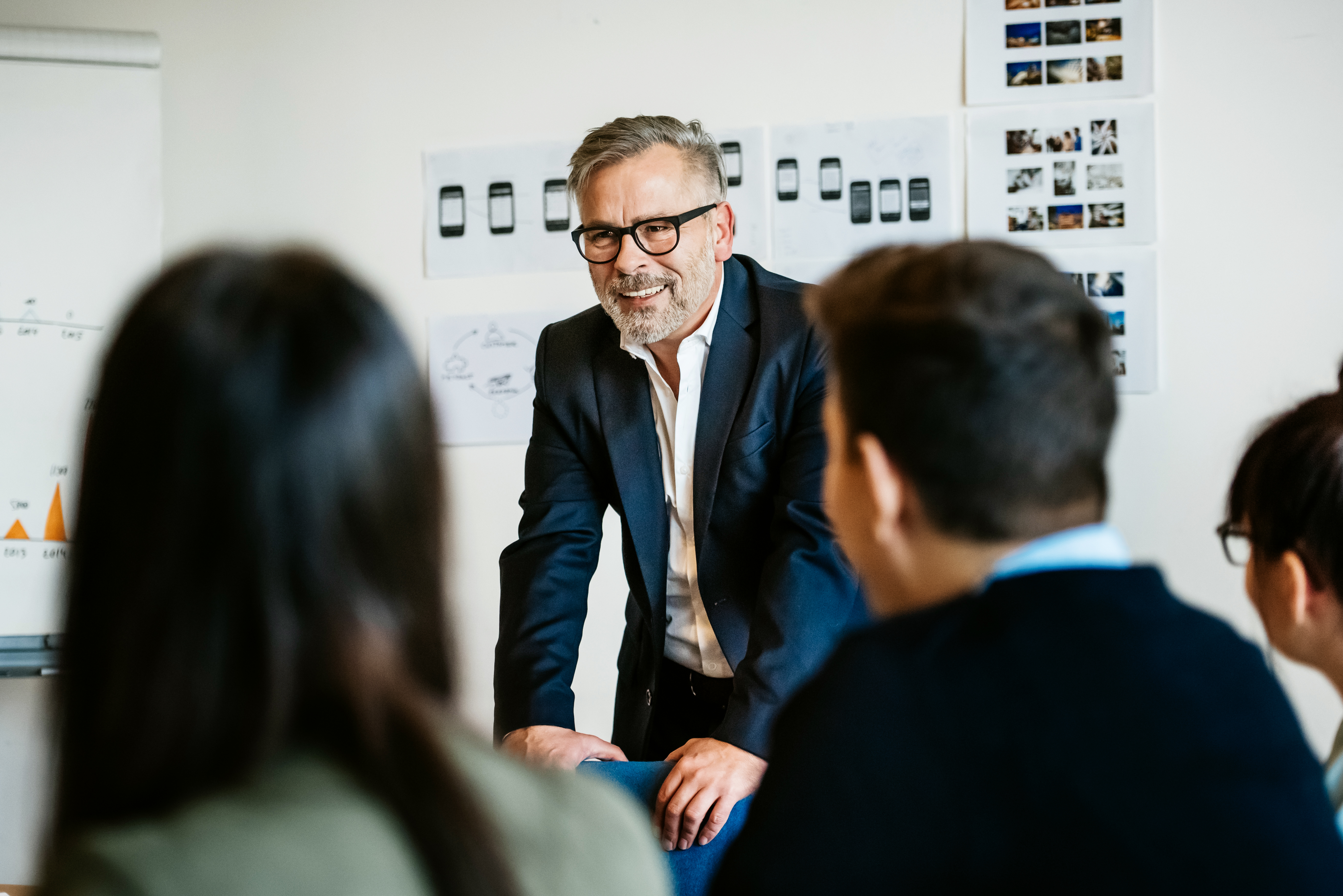 A man in a suit and glasses leans on a table, smiling, during a meeting. He is surrounded by seated people, with design sketches and photos on the wall behind him.