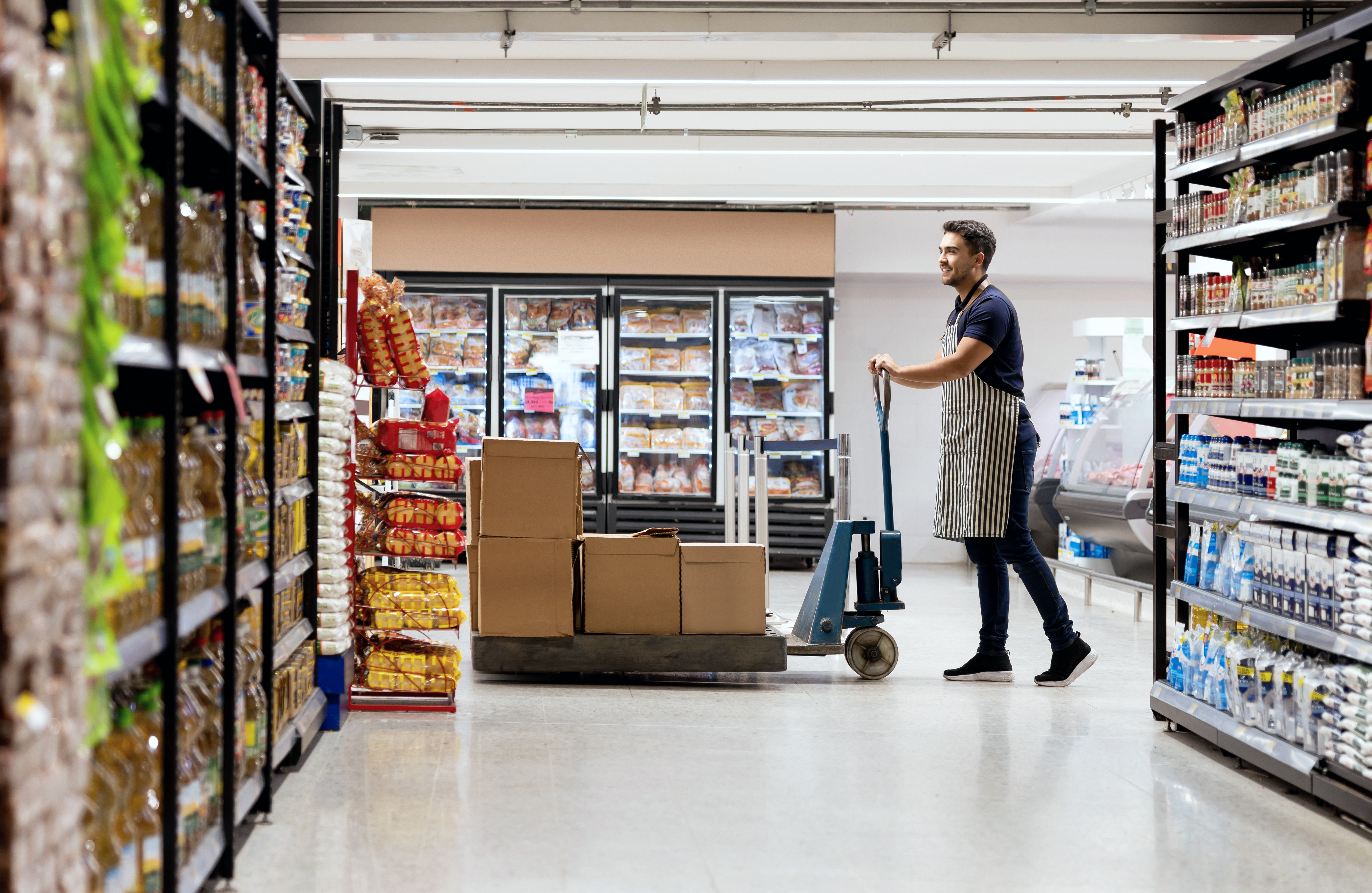 A supermarket worker uses a hand truck to transport boxes down an aisle, flanked by shelves and refrigerated display cases.