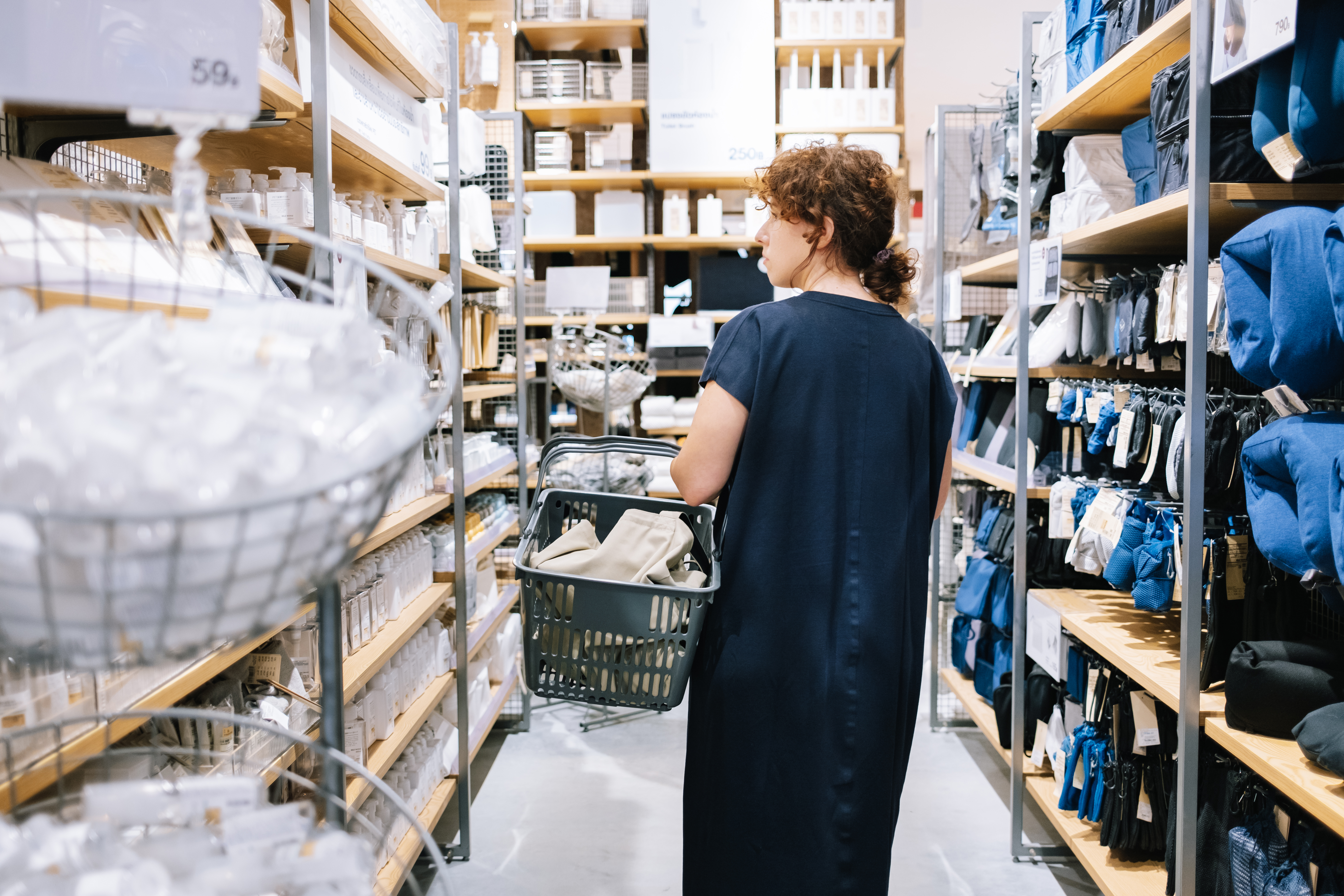 Person in a navy dress holding a shopping basket, browsing shelves stocked with household items in a store.