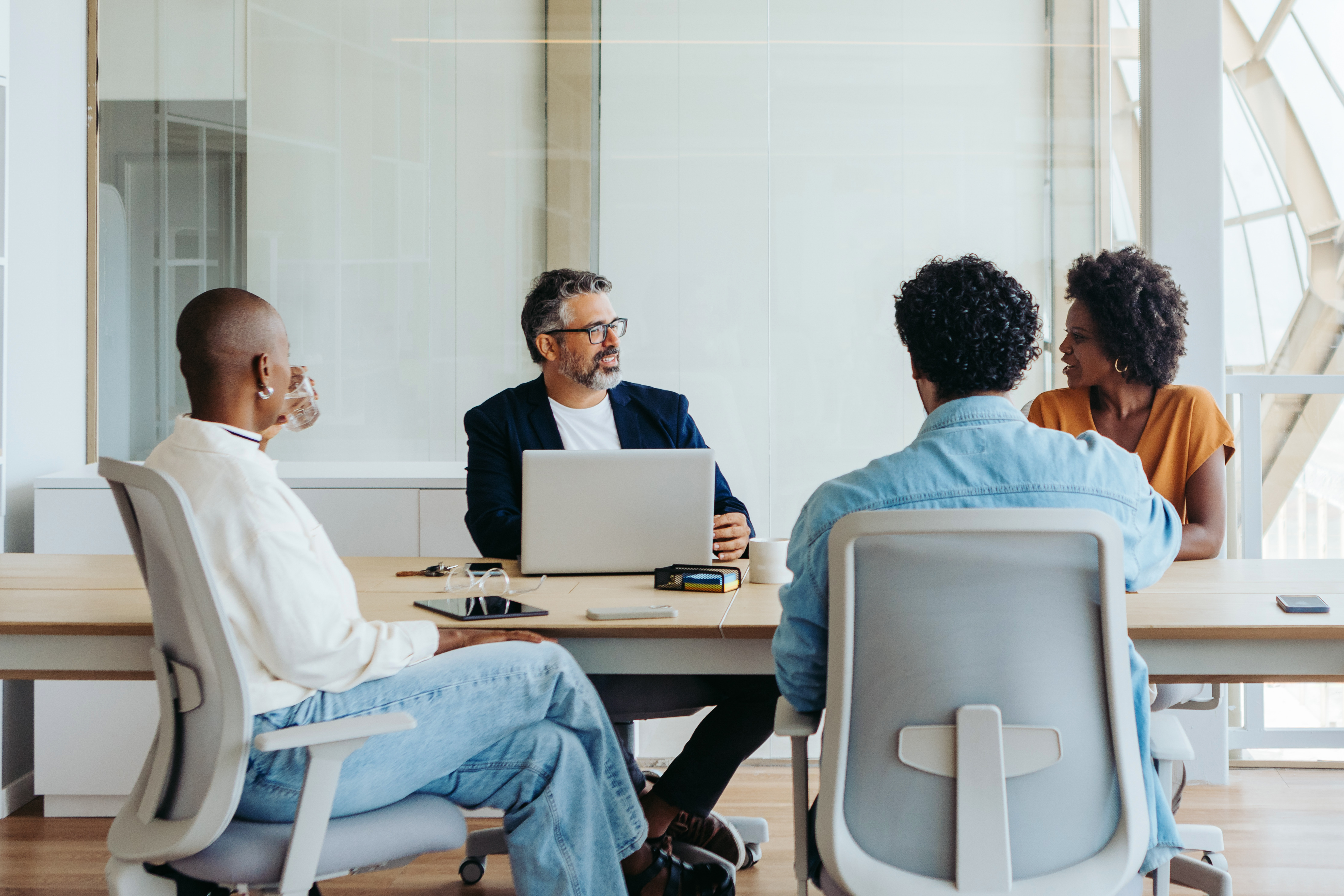 A diverse group sits around a modern conference table, engaged in discussion, with a laptop and documents displayed.