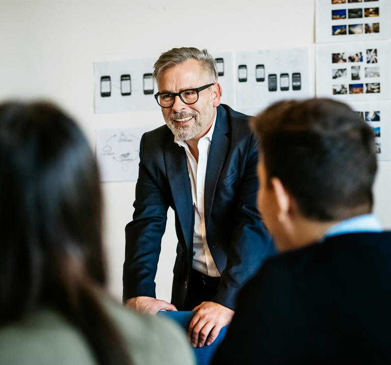 A man in a suit and glasses leans on a table, smiling, during a meeting. He is surrounded by seated people, with design sketches and photos on the wall behind him.