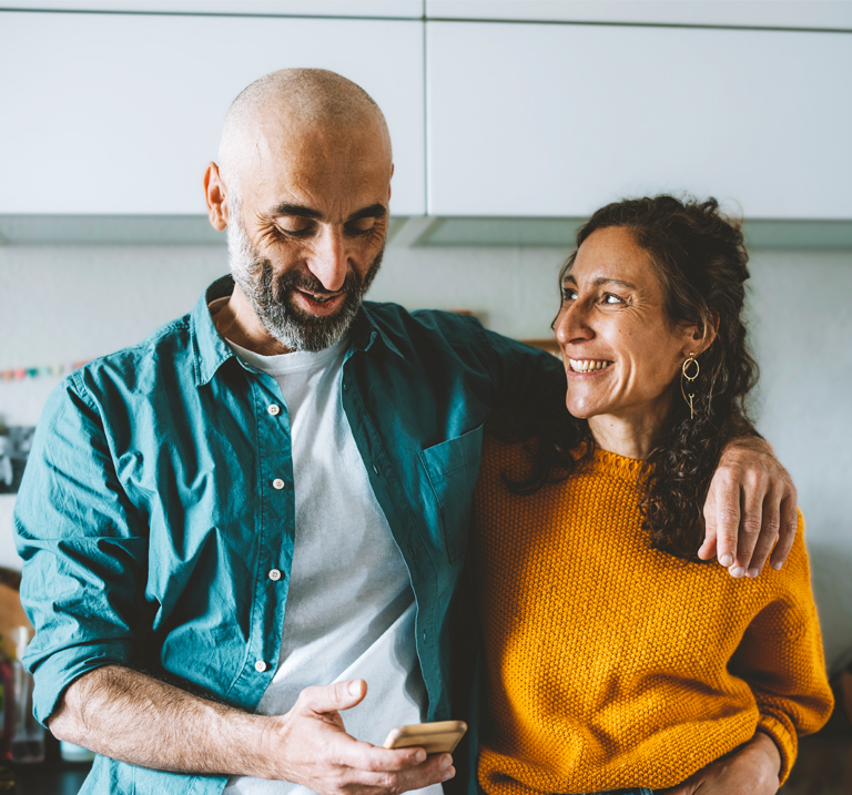 A couple stands close together, smiling, with one holding a phone, in a bright kitchen setting with modern decor.