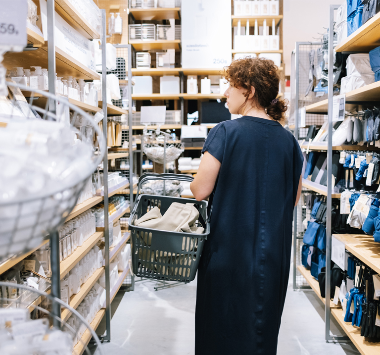 Person in a navy dress holding a shopping basket, browsing shelves stocked with household items in a store.