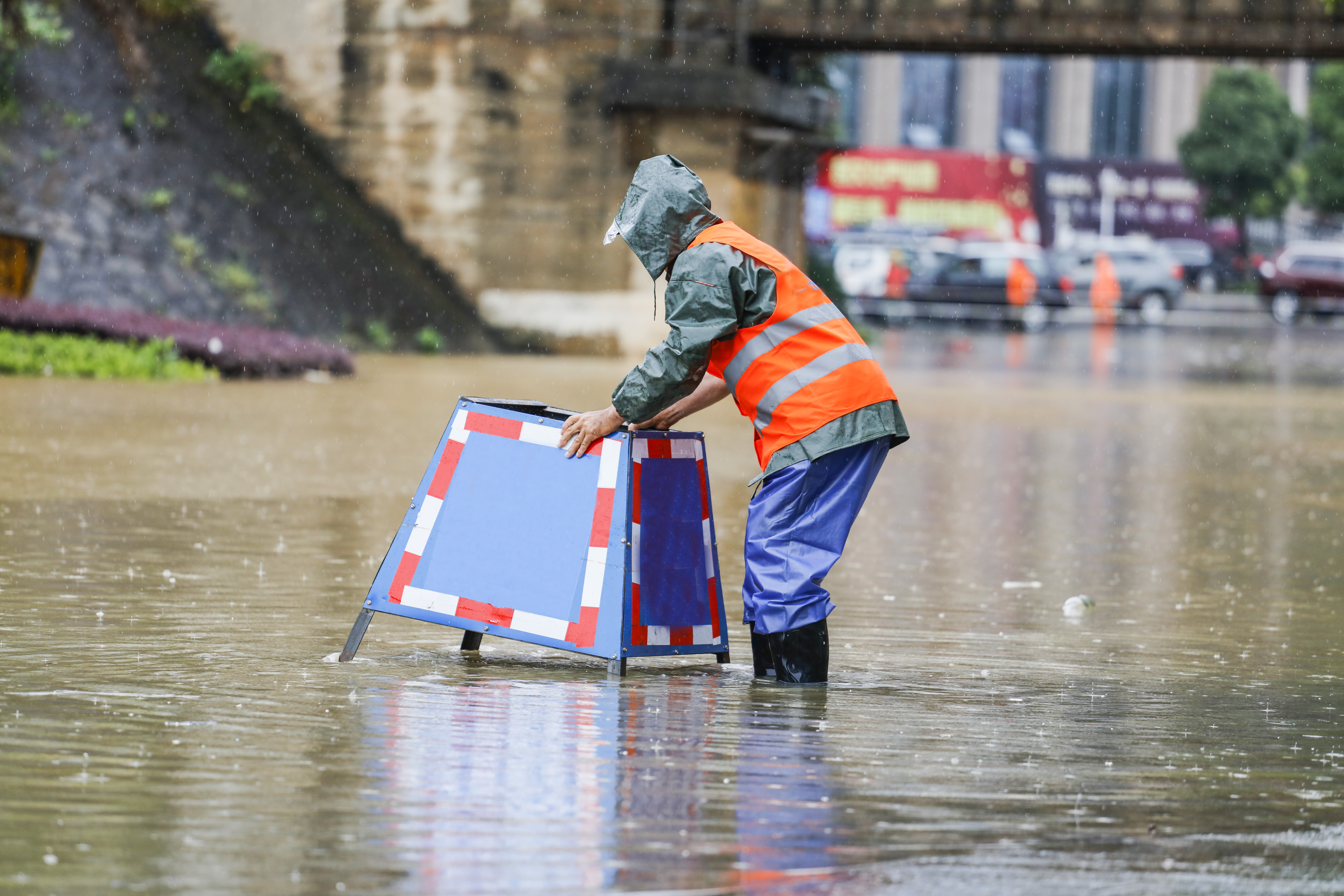 A worker in waterproof gear sets up a warning sign in a flooded street, heavy rain falling around him.