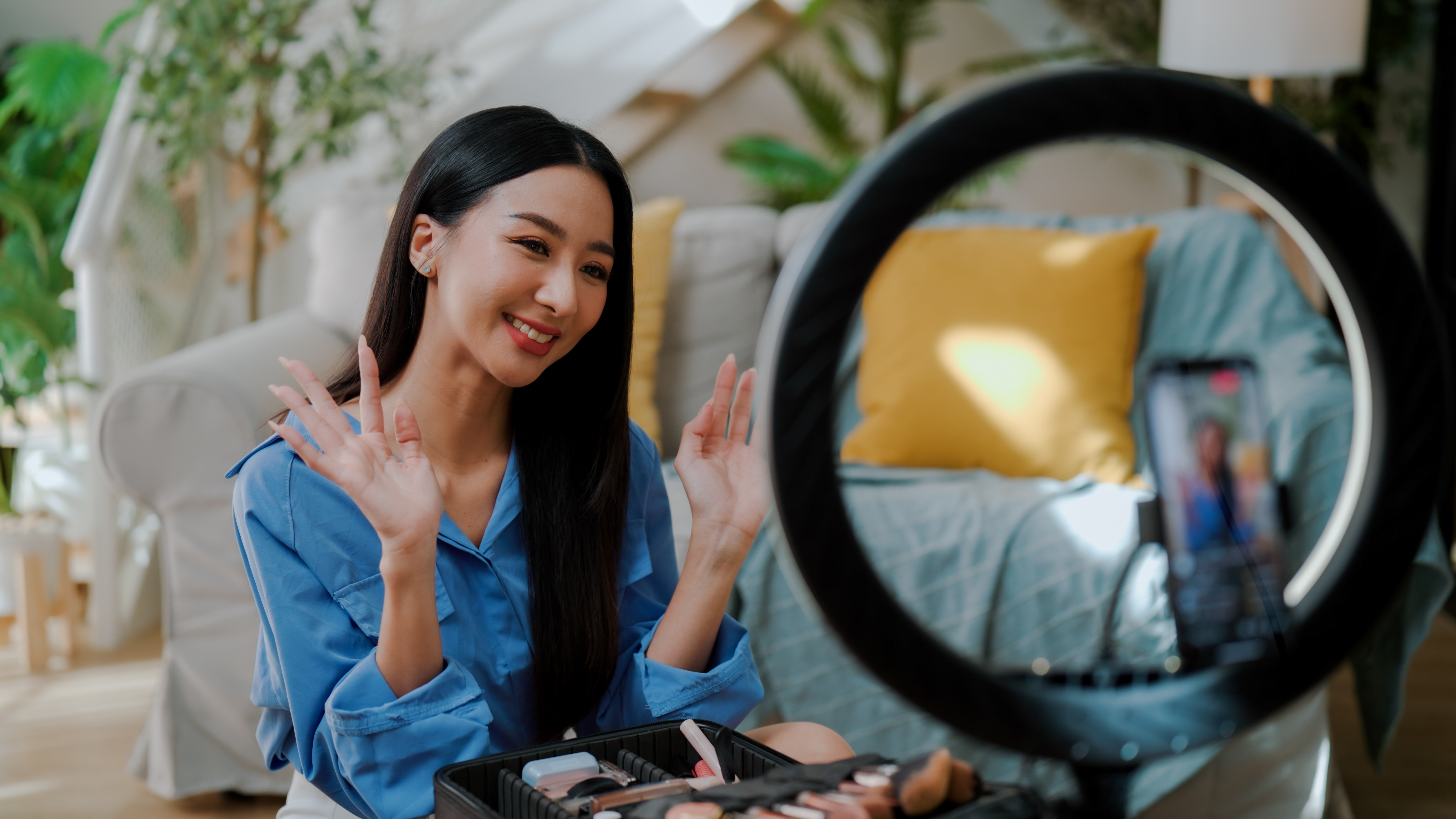 Person in a blue shirt faces a ring light, smiling with open hands. A makeup kit is visible in front. Vibrant living room setting.